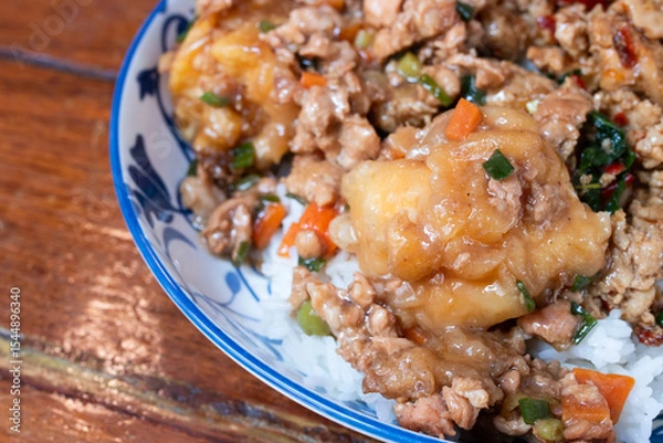 Fototapeta A Thai rice dish topped with stir-fried minced pork, vegetables, and battered fried egg, served in a patterned ceramic bowl on a wooden table.