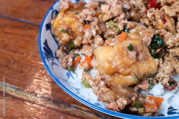 Fototapeta A Thai rice dish topped with stir-fried minced pork, vegetables, and battered fried egg, served in a patterned ceramic bowl on a wooden table.