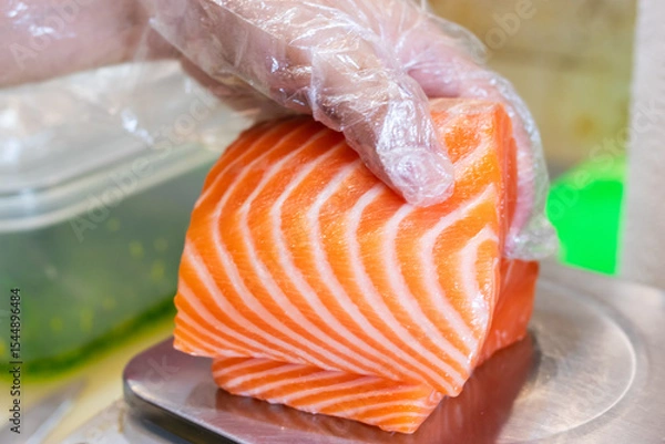 Fototapeta Fresh raw salmon fillet being held with a gloved hand on a stainless steel scale, ready for preparation in sushi or sashimi dishes.