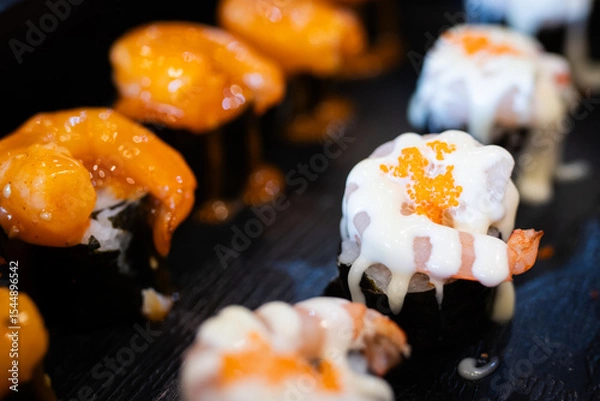 Fototapeta Assorted sushi topped with creamy sauces and shrimp, served on a black plate, highlighting the vibrant textures and savory presentation.