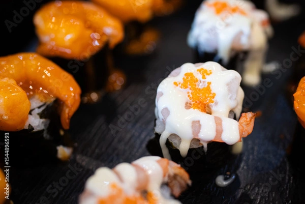 Fototapeta Assorted sushi topped with creamy sauces and shrimp, served on a black plate, highlighting the vibrant textures and savory presentation.