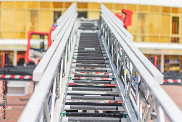 Fototapeta Red ladder truck with safety handrails, extended aerial device, multiple compartments, daylight lighting, straightforward style