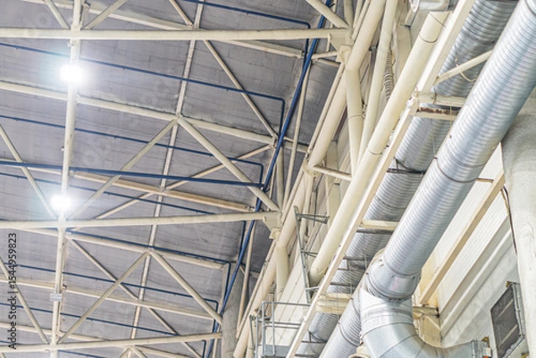 Fototapeta Industrial space with large open room, high ceilings supported by metal beams and pillars, network of multi-colored pipes, artificial lighting, concrete floor, documentary photography style, perspect