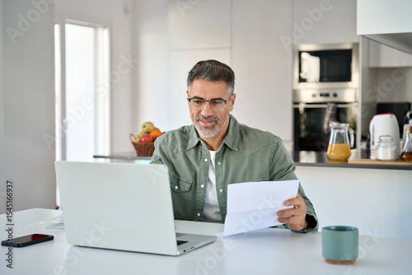 Obraz Middle aged mature man senior customer wearing glasses looking at laptop computer holding paper bill checking financial invoice or tax document making online banking payments working at home table.