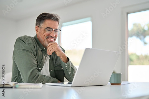 Obraz Smiling wearing eyeglasses looking at computer sitting at table. Happy older mature middle aged man using laptop hybrid working online, elearning, browsing web, searching online at home.