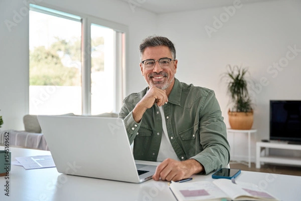 Obraz Happy smiling mature middle aged older professional man wearing eyeglasses looking at camera sitting at living room table, using laptop computer working online at casual home. Portrait.