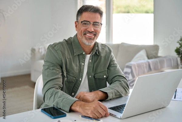 Obraz Portrait of happy smiling mature middle aged older professional man wearing glasses looking at camera sitting at living room table, using laptop computer working online at casual home.