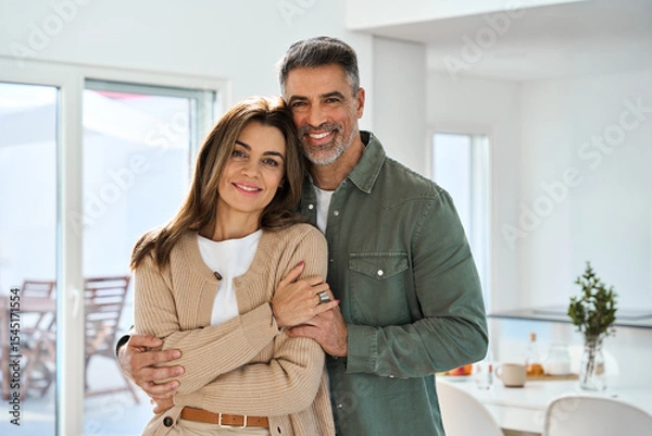 Obraz Smiling affectionate loving middle aged couple, happy romantic mature older Latin man and woman in their 50s hugging looking at camera standing together in modern kitchen at home. Portrait.