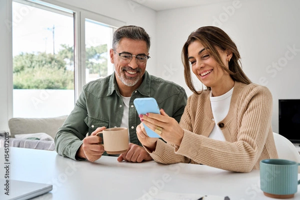 Obraz Smiling mature older man and woman holding cellphone browsing internet, texting message on mobile cell phone technology. Middle aged happy couple using smartphone relaxing at living room table at home