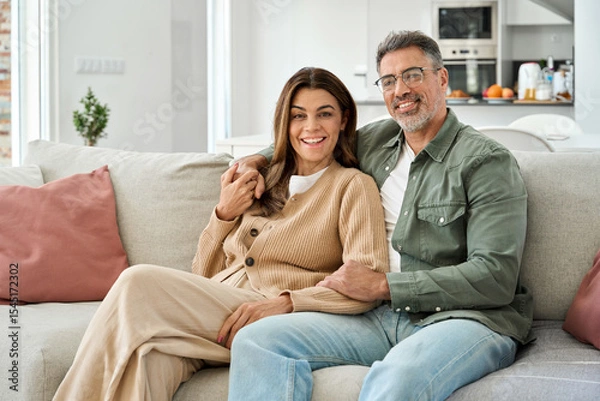 Obraz Family portrait of happy middle aged mature romantic couple in their 50s hugging sitting on couch at home. Smiling older man and woman in love relaxing on sofa together in house living room.