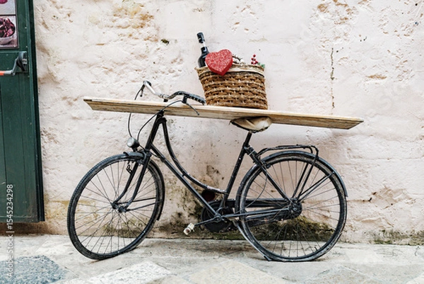 Fototapeta Matera, Italy. An old bicycle leans against a stone wall in Sassi di Matera, Italy, with a basket holding wine bottle and a red heart, used as a charming display or rustic street decor. 