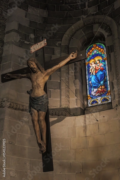 Fototapeta Matera, Italy. Wooden crucifix of Jesus in the Church of San Giovanni Battista, Sassi of Matera. A stained glass window depicting the Madonna and Child glows in the background. Vertical. 
