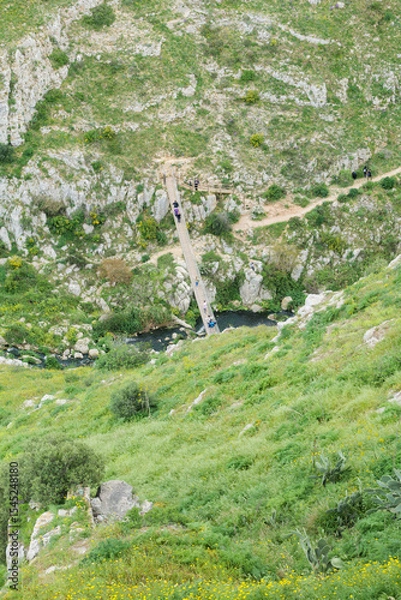 Fototapeta Matera, Italy. Scenic view of the Gravina river below Matera's Sassi. People cross a Tibetan bridge along path 406 in the Murgia Materana Park, surrounded by rocky green hills. Vertical image.
