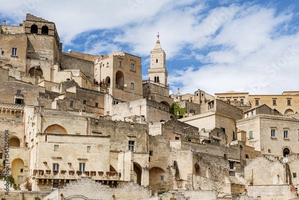 Fototapeta Matera, Italy. Scenic view of the historic center of the Sassi di Matera, Italy, with ancient stone houses and the bell tower of Matera Cathedral under a bright blue sky.