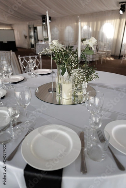 Fototapeta Wedding reception table beautifully arranged with white roses and candles atop a mirror. Elegant tableware includes classic plates and glassware, creating a sophisticated atmosphere.