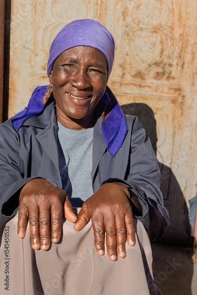 Fototapeta village single old african woman with headscarf sited in front of the house , in the yard, close portrait