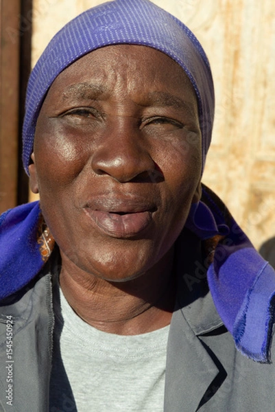 Fototapeta village single old african woman with headscarf sited in front of the house , in the yard, close portrait