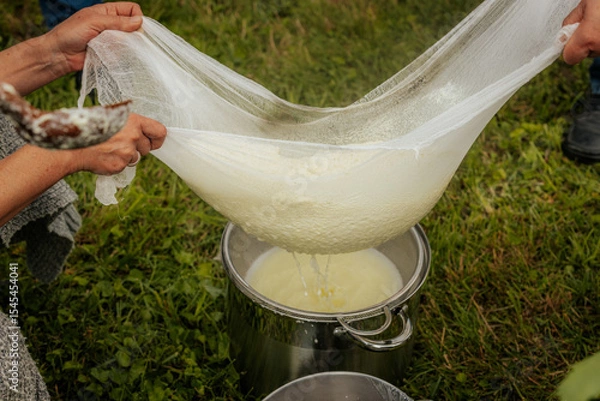 Fototapeta Fresh cheese is being strained through cloth, held by people outdoors using large green leaves, with whey dripping into a container below.