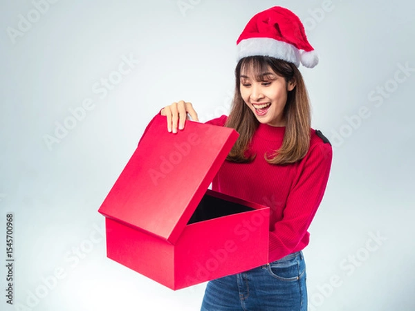 Fototapeta Asian woman in red sweater and Santa hat looking surprised while opening a large red gift box, with joyful expression and festive spirit on a plain light background