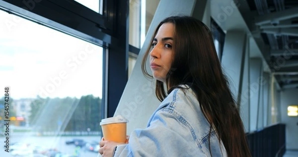 Obraz Young woman wearing a denim jacket stands in an airport terminal, holding a takeaway coffee cup and checking the time on her smartwatch while looking out the large window at the planes and runway