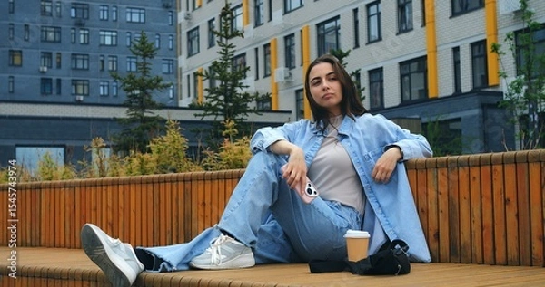 Obraz Modern buildings provide the backdrop as a young woman, dressed in casual attire, enjoys a beverage while engaging with her smartphone on a comfortable bench