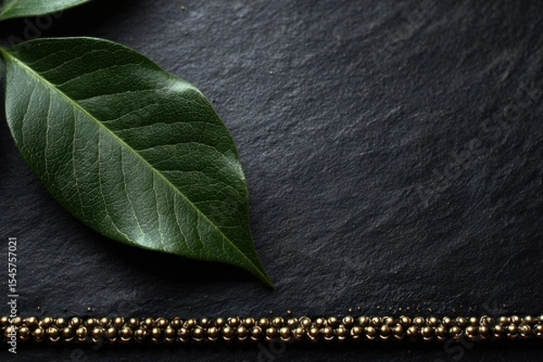 Fototapeta A green leaf on a black surface next to a gold chain
