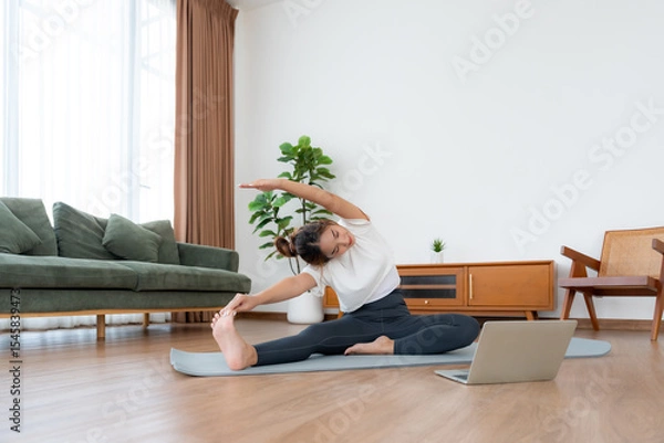 Fototapeta Young Asian woman stretching yoga workout on exercise mat while online training class with computer laptop in living room