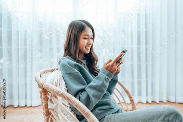 Fototapeta Young Asian Woman Relaxing in Modern Wicker Chair Near Window with Sheer Curtain, Happy Female Using Mobile Smartphone Enjoying at Home, Comfort and Cozy