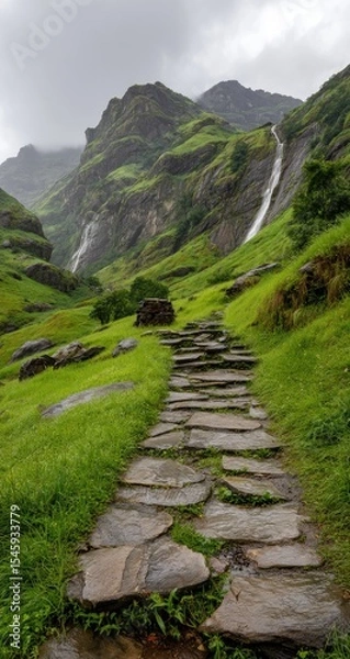 Fototapeta Stone path ascends verdant hillside towards cascading waterfalls nestled within imposing, mist-shrouded mountains