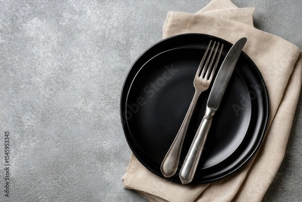 Fototapeta Two black plates, silver fork and knife rest on a beige linen napkin atop a grey textured surface