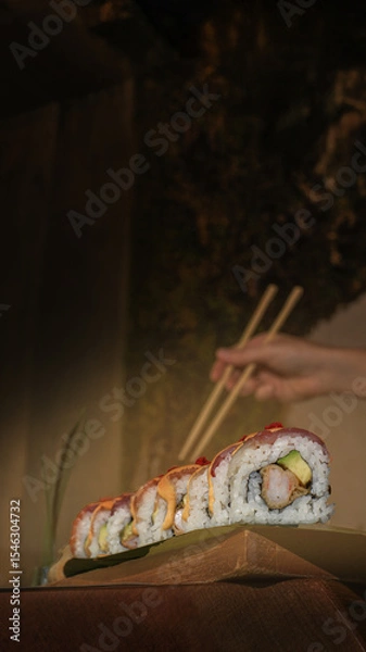 Fototapeta Hand holding sushi roll with chopsticks, showcasing fresh ingredients. Vertical orientation