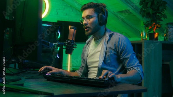 Fototapeta Emotional angry mad furious talking in microphone video game loss addiction Caucasian guy male man streamer gamer wearing headphones playing virtual battle lost fail internet computer at night home