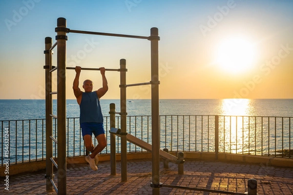 Obraz A senior man does pull-ups on a bar by the sea at sunrise. His silhouette highlights calmness, vitality, and mindful aging. Golden backlight enhances the peaceful morning atmosphere.