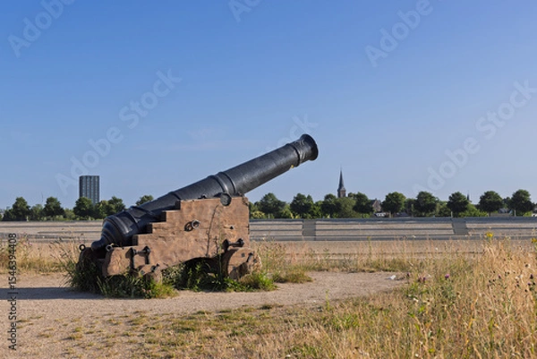 Obraz An historic black cannon, a remnant of past military might, stands in a dry, grassy field under a clear blue sky, with a distant church spire and modern buildings.