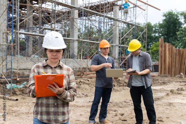Obraz Asian female engineer writing on clipboard while two male workers review plans on digital devices at a construction site. Teamwork and supervision in civil engineering project.