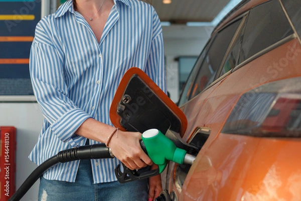 Fototapeta A person holds a fuel nozzle, refueling an orange vehicle at a gas station during the afternoon. The atmosphere is busy and practical.