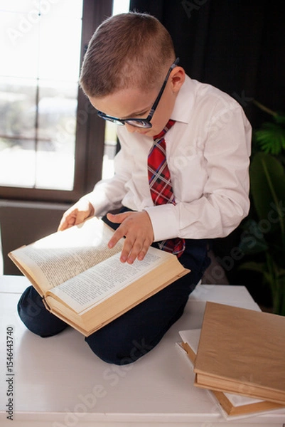 Fototapeta A 7-year-old child in glasses and  dressed school uniform poses at the school table indoor/ The boy learn the book