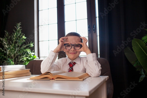 Fototapeta A 7-year-old child in glasses and  dressed school uniform poses at the school table indoor/ The boy learn the book