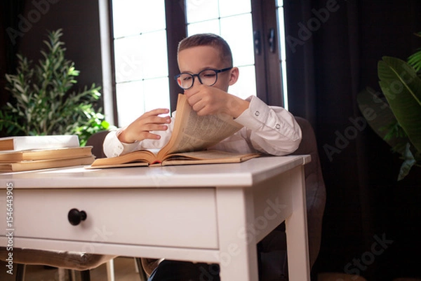 Fototapeta A 7-year-old child in glasses and  dressed school uniform poses at the school table indoor/ The boy learn the book