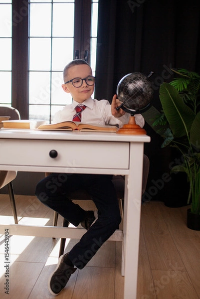 Fototapeta A 7-year-old child in glasses and  dressed school uniform poses at the school table indoor/ The boy learn the book