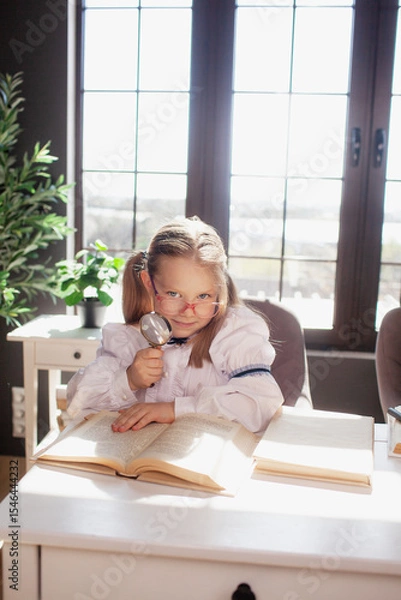 Fototapeta A 7-year-old child in glasses dressed in school uniform poses at the school table indoor. The girl holds globe