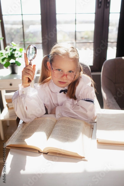 Fototapeta A 7-year-old child in glasses dressed in school uniform poses at the school table indoor. The girl holds globe
