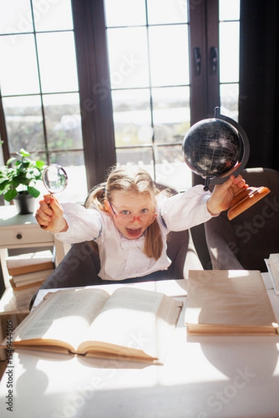 Fototapeta A 7-year-old child in glasses dressed in school uniform poses at the school table indoor. The girl holds globe