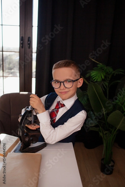 Fototapeta A 7-year-old child in glasses and dressed school uniform poses at the school table indoor. The boy looks to the microscope