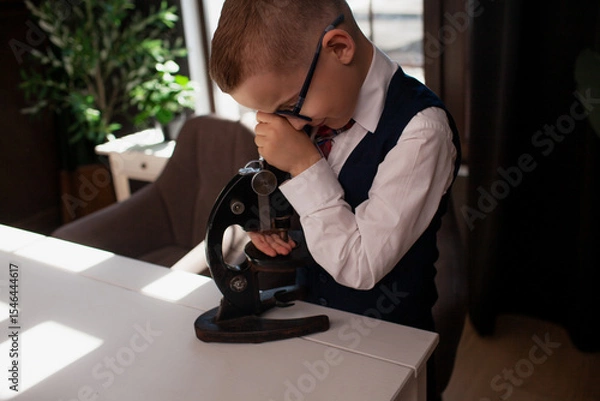 Fototapeta A 7-year-old child in glasses and dressed school uniform poses at the school table indoor. The boy looks to the microscope