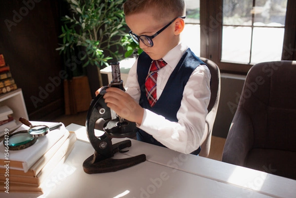 Fototapeta A 7-year-old child in glasses and dressed school uniform poses at the school table indoor. The boy looks to the microscope