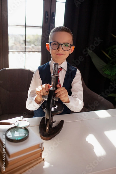 Fototapeta A 7-year-old child in glasses and dressed school uniform poses at the school table indoor. The boy looks to the microscope