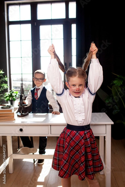 Fototapeta A 7-year-old child in glasses dressed in  school uniform poses at the school table indoor The boy and girl looks to the microscope
