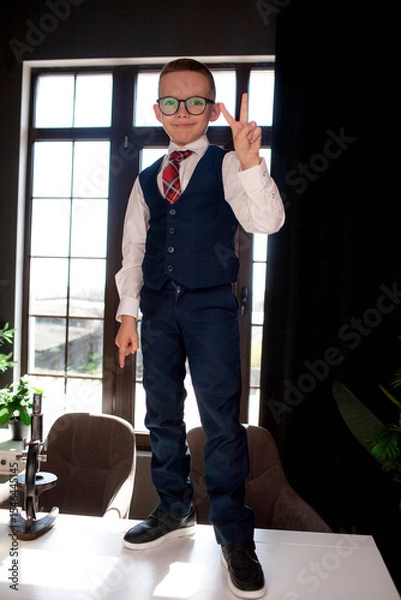 Fototapeta A 7-year-old child in glasses and dressed  school uniform poses at the school table indoor
