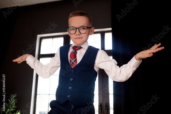 Fototapeta A 7-year-old child in glasses and dressed  school uniform poses at the school table indoor